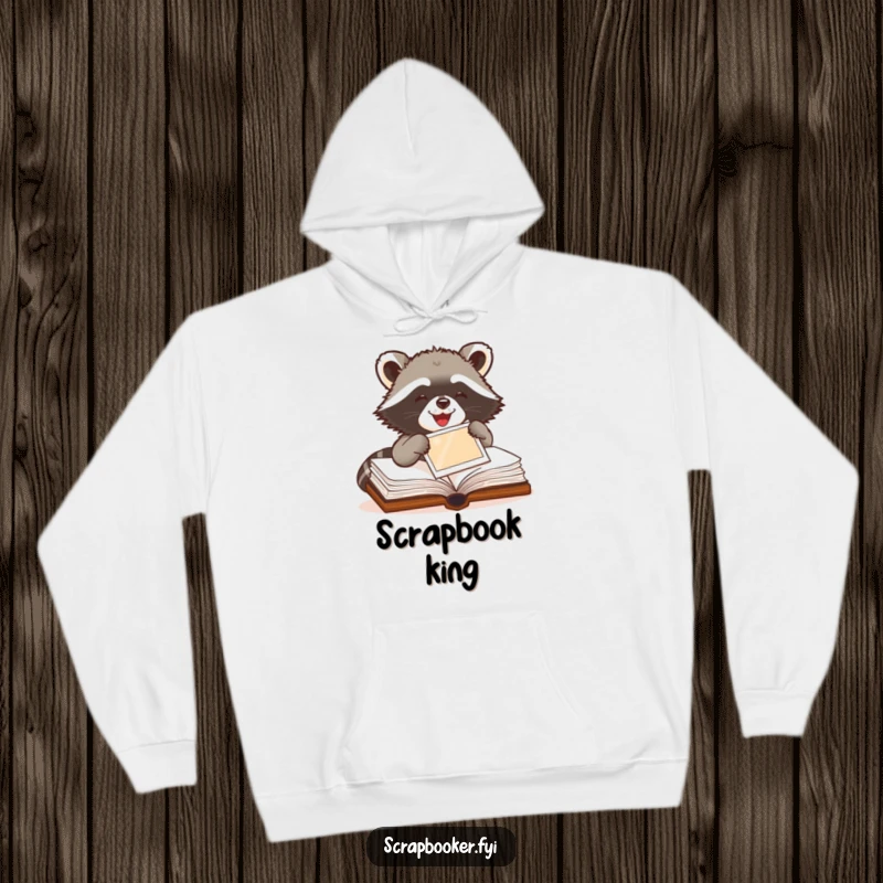 Funny Raccoon Hoodie depicting a delighted raccoon meticulously pasting a photo into a scrapbook, great for warmth and humor.